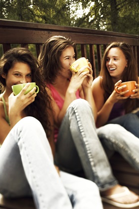 three_relaxed_teen_girls_drinking_coffee_sitting_on_balcony_on_forest_background